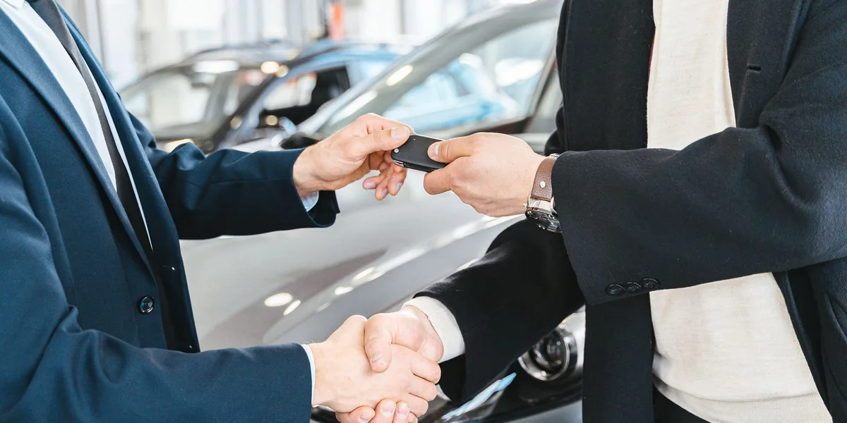 Two people shake hands as one passes a black car key fob to the other, with a silver car out of focus behind them.
