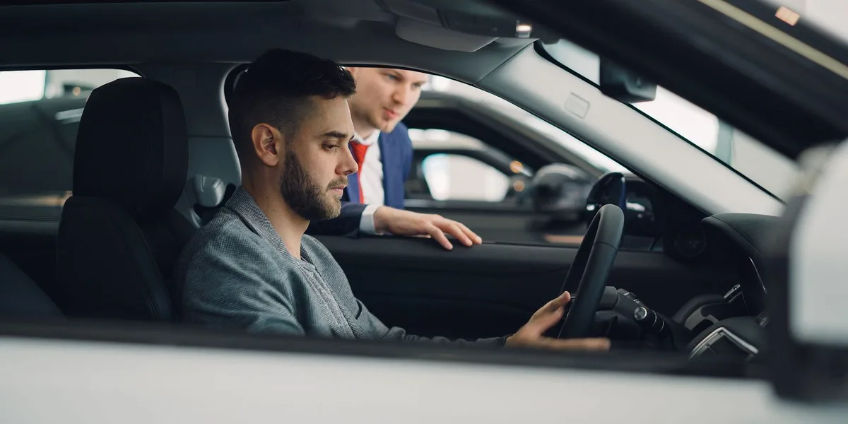 A buyer sits in the driver's seat of a used car with his hands near the wheel while a salesperson leans in at the open window to answer a question.
